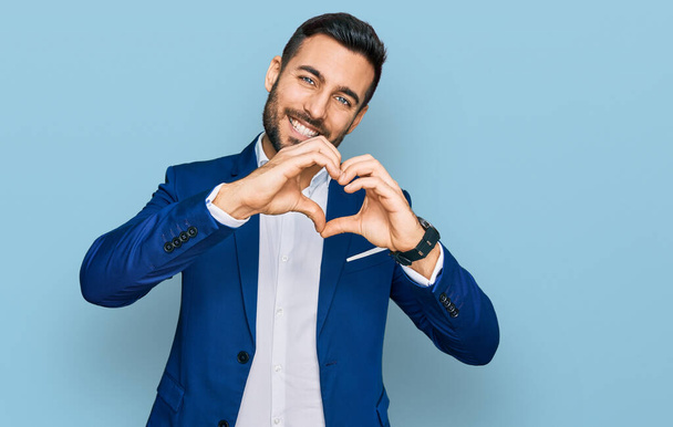 Jovem hispânico vestindo jaqueta de negócios sorrindo no amor fazendo forma de símbolo do coração com as mãos. conceito romântico.  - Foto, Imagem
