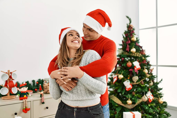Jeune couple étreignant et souriant heureux portant un chapeau de Noël à la maison. - Photo, image