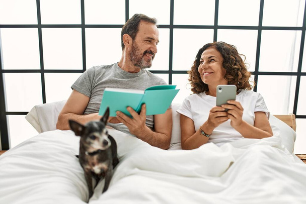 Medioevo coppia ispanica lettura libro e utilizzando smartphone. Sdraiato sul letto con cane a casa. - Foto, immagini