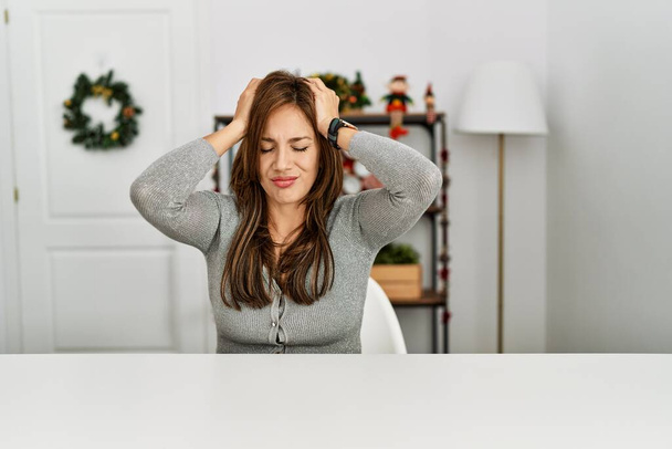 Mujer latina joven sentada en la mesa por la decoración de Navidad que sufre de dolor de cabeza desesperada y estresada porque el dolor y la migraña. manos en la cabeza.  - Foto, imagen