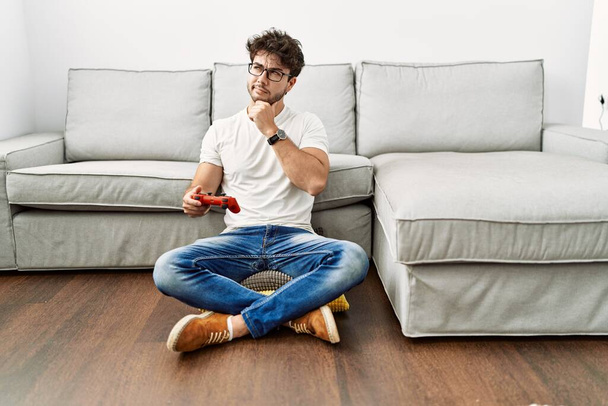 Hispanic man playing video game holding controller by the sofa serious face thinking about question with hand on chin, thoughtful about confusing idea  - Foto, imagen