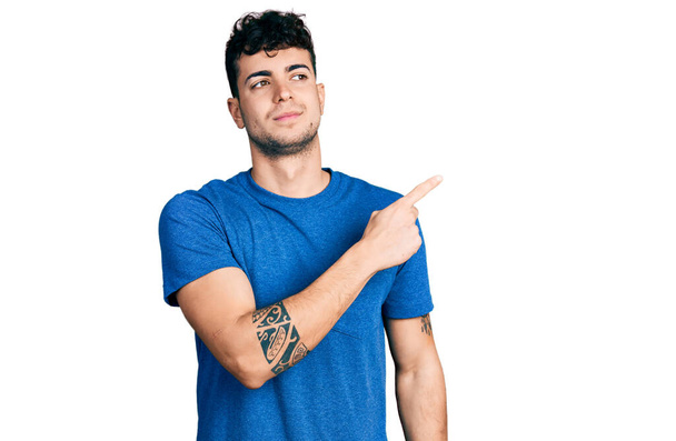 Joven hombre hispano con camiseta casual señalando con el dedo de la mano a un lado mostrando publicidad, cara seria y tranquila  - Foto, imagen