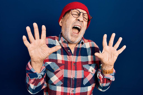 Knappe volwassen man draagt hipster look met wollen pet gek en gek schreeuwen en schreeuwen met agressieve uitdrukking en armen omhoog. frustratieconcept.  - Foto, afbeelding