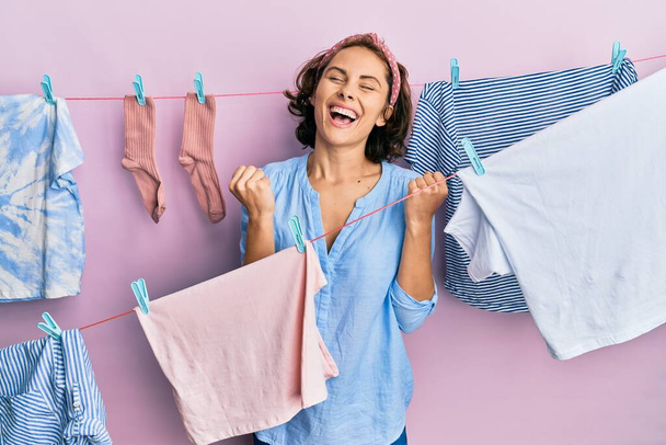 Young brunette woman doing laundry around string hangs very happy and excited doing winner gesture with arms raised, smiling and screaming for success. celebration concept.  - Fotografie, Obrázek