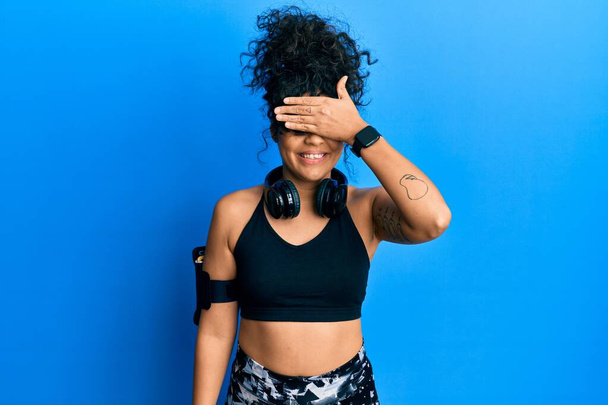 Young hispanic woman wearing gym clothes and using headphones smiling and laughing with hand on face covering eyes for surprise. blind concept.  - Foto, immagini