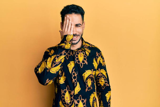 Young hispanic man wearing casual shirt covering one eye with hand, confident smile on face and surprise emotion.  - Photo, Image