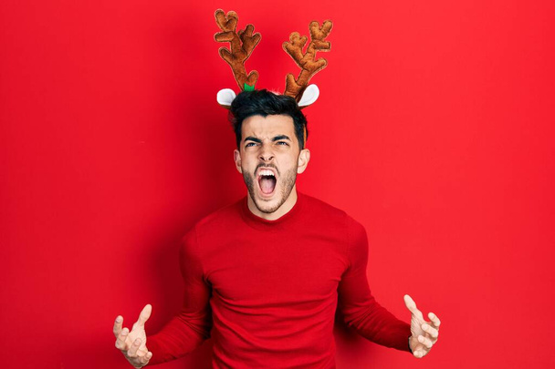 Νεαρός Ισπανός που φοράει χαριτωμένα χριστουγεννιάτικα κέρατα ταράνδου τρελός και τρελός φωνάζοντας και φωνάζοντας με επιθετική έκφραση και χέρια σηκωμένα. έννοια απογοήτευσης.  - Φωτογραφία, εικόνα