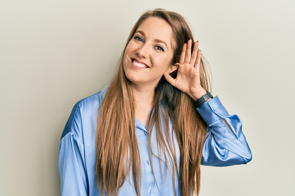 Egy fiatal szőke nő alkalmi kék ingben, aki fültől fülig mosolyog, miközben egy pletykát vagy pletykát hallgat. süketség fogalma.  - Fotó, kép