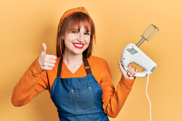 Redhead young woman holding pastry blender electric mixer smiling happy and positive, thumb up doing excellent and approval sign  - Fotografie, Obrázek