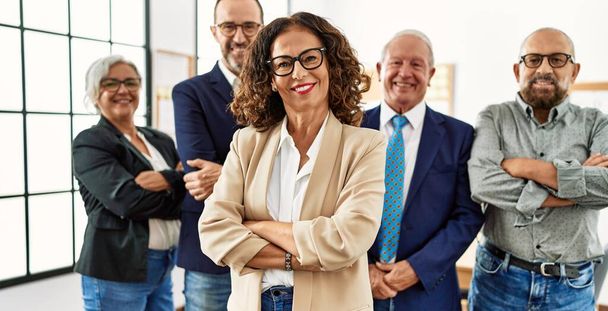 Группа бизнесменов среднего возраста улыбается счастливой стоя со скрещенными руками жестом в офисе - Фото, изображение
