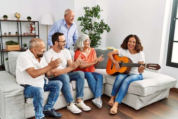 Ομάδα μεσήλικων φίλων που κάνουν πάρτι παίζοντας κλασική κιθάρα καθισμένοι στον καναπέ στο σπίτι. - Φωτογραφία, εικόνα