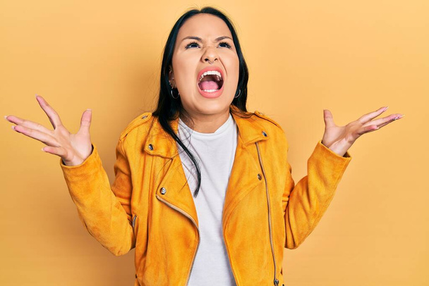 黄色の革のジャケットを身に着けてピアス鼻を持つ美しいヒスパニック系の女性は狂気と怒りの叫びと積極的な表現と腕を上げて叫んでいます。フラストレーションコンセプト.  - 写真・画像