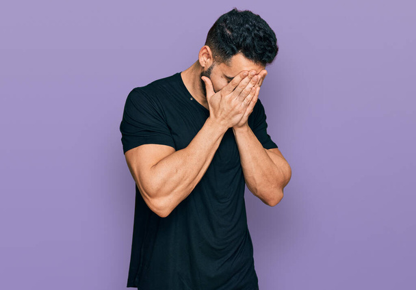 Spaanse man met baard draagt casual zwart t-shirt met droevige uitdrukking bedekken gezicht met handen tijdens het huilen. depressie concept.  - Foto, afbeelding