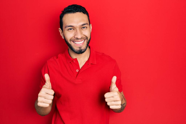 Hispanischer Mann mit Bart trägt legeres rotes T-Shirt und macht positive Geste mit der Hand, Daumen hoch lächelnd und glücklich. Fröhlicher Ausdruck und Siegergeste.  - Foto, Bild