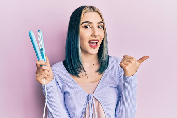Young modern girl holding hair straightener pointing thumb up to the side smiling happy with open mouth  - Photo, Image