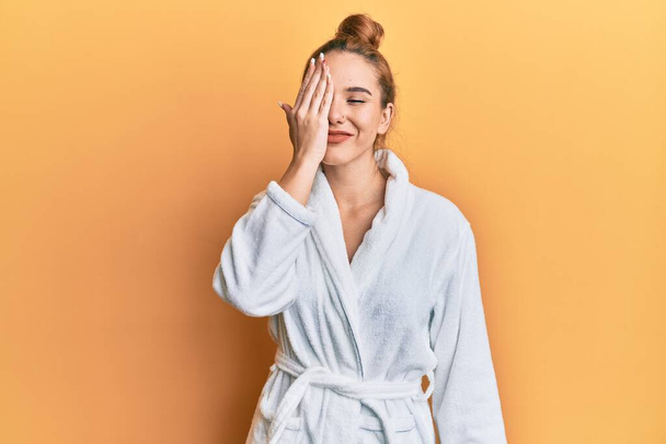 Young blonde woman wearing bathrobe covering one eye with hand, confident smile on face and surprise emotion.  - Foto, Imagen