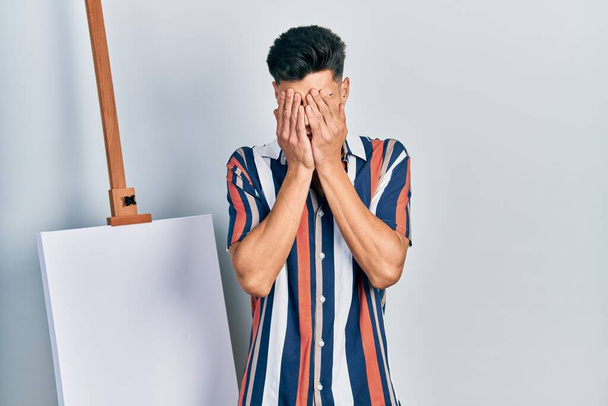 Young hispanic man standing close to empty canvas with sad expression covering face with hands while crying. depression concept.  - Fotoğraf, Görsel