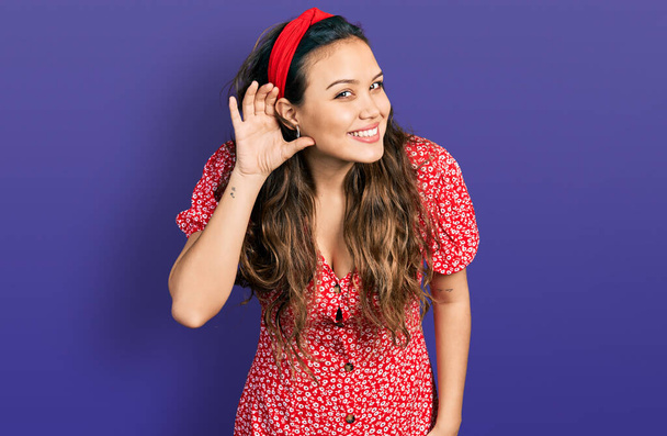 Молода іспаномовна дівчина в повсякденному одязі посміхається рукою через вухо слухаючи чутки або плітки. концепція глухоти
.  - Фото, зображення