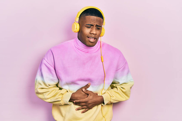 Fiatal fekete férfi zenét hallgat fülhallgatóval a hasán, mert hányinger, fájdalmas betegség rossz közérzet. fájdalom koncepció.  - Fotó, kép