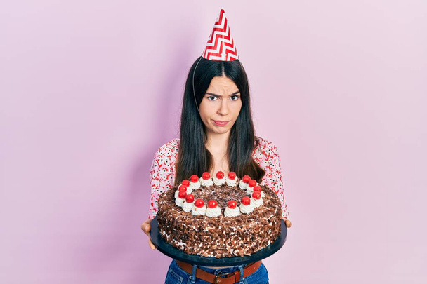 Молода брюнетка, яка святкує день народження, тримає великий шоколадний торт скептично і нервово, розчаровується через проблеми. негативна людина
.  - Фото, зображення