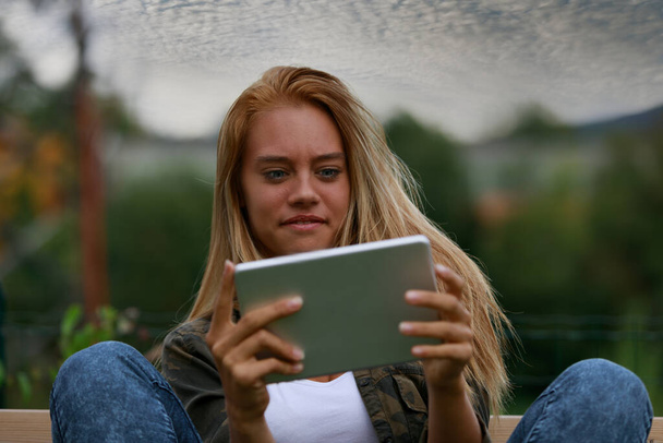 Zszokowana młoda kobieta patrząc na tablet w sceptycyzmie z szerokimi oczami i grymasem, relaksuje się czytając lub oglądając media na zewnątrz o zmierzchu - Zdjęcie, obraz