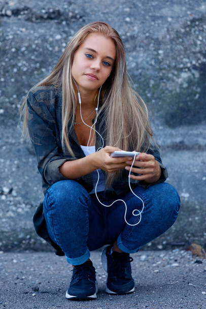 Jeune femme blonde à la mode accroupie pour écouter sa musique sur un téléphone portable et des écouteurs regardant attentivement la caméra avec un sourire calme - Photo, image