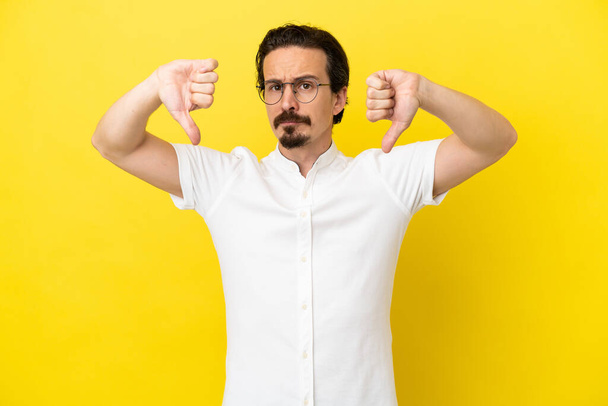 Young caucasian man isolated on yellow background showing thumb down with two hands - Photo, Image