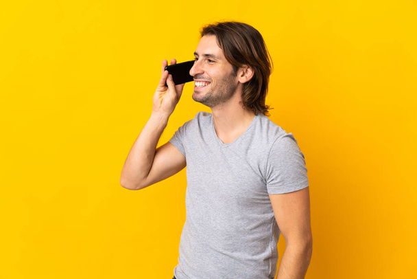 Young handsome man isolated on yellow background keeping a conversation with the mobile phone - Фото, изображение