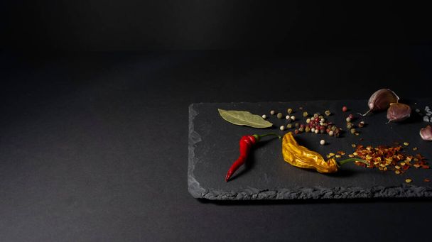 Spices on a stone plate. Black background. Black stone. Garlic, sea salt, pepper, chili, hot pepper, bay leaf - Фото, изображение