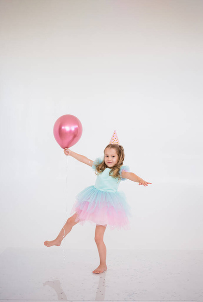 una niña rubia en un vestido elegante baila con un globo rosa sobre un fondo blanco con un lugar para el texto - Foto, Imagen