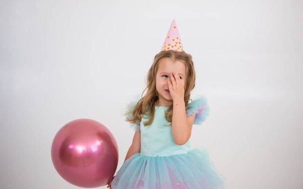 a happy little girl in a paper cap and a smart dress stands with a pink balloon on a white background with a place for text - Photo, Image