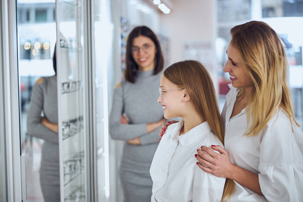 Hermosa rubia mujer caucásica y chica de la escuela mirando al espejo en la tienda de oftalmología - Foto, imagen