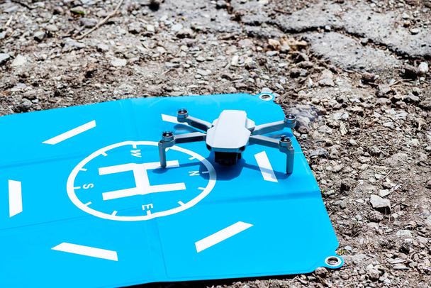 Το drone στέκεται σε ένα μπλε πεδίο προσγείωσης σε βραχώδη, οι πλαστικές προπέλες είναι ενεργοποιημένες και η κάμερα στο gimbal είναι ορατή. - Φωτογραφία, εικόνα