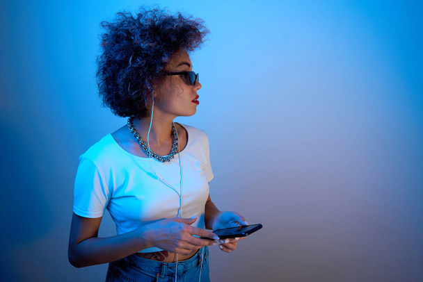 Trendy girl with afro curls uses a smartphone and headphones on a studio background with copy space - Фото, изображение