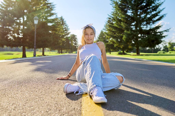 Fashionable trendy teenage hipster female sitting on road in park - Foto, afbeelding
