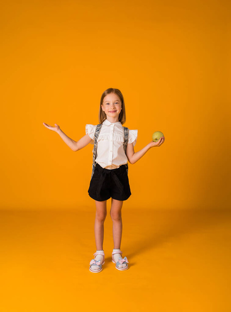 a little schoolgirl in a uniform and with a backpack stands and holds an apple on a yellow background with a copy of the space - Photo, Image
