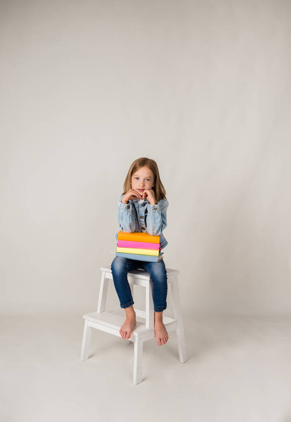 una niña rubia en denim está sentada en una silla con libros de texto sobre un fondo blanco con una copia del espacio - Foto, imagen