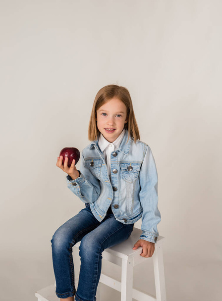 una piccola ragazza bionda in denim tiene una mela rossa su uno sfondo bianco con un posto per il testo - Foto, immagini