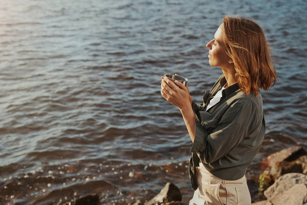 Kadın elinde çay fincanıyla kıyıda duruyor. - Fotoğraf, Görsel
