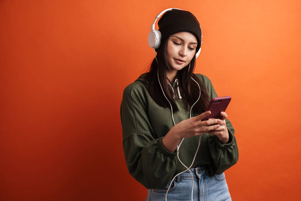 Happy young casual brunette woman with long hair wearing hat standing over red background, wearing headphones, listening to music, holding mobile phone - Φωτογραφία, εικόνα