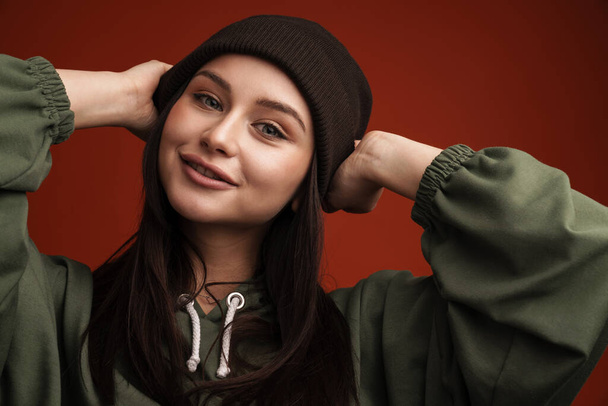 Happy young casual brunette woman with long hair wearing hat standing over red background - Φωτογραφία, εικόνα