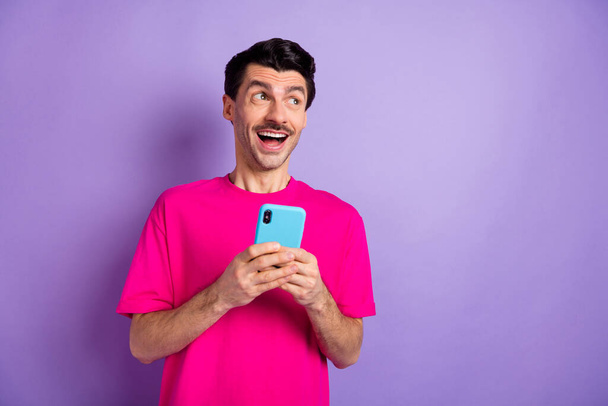 Photo of attractive impressed young man wear pink t-shirt holding modern gadget looking empty space isolated purple color background - Foto, Bild