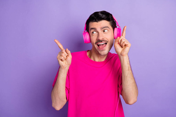 Photo of sweet funky young man wear pink t-shirt listening music headphones looking empty space isolated purple color background - Photo, image