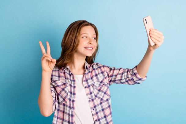 Photo of cute sweet school girl wear plaid shirt showing v-sign tacking selfie modern gadget isolated blue color background - Foto, Bild