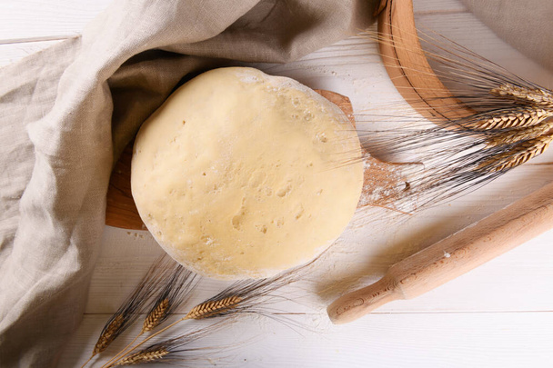 Сырое домашнее тесто для хлеба или пиццы с колосьями пшеницы, льняной салфетки и муки на столе - Фото, изображение