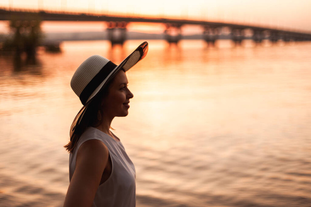 A woman at the bridge over the river at sunset. Profile portrait of a young woman in a straw hat admiring the sunset on the beach of a river with a bridge on a summer evening - Foto, Imagem