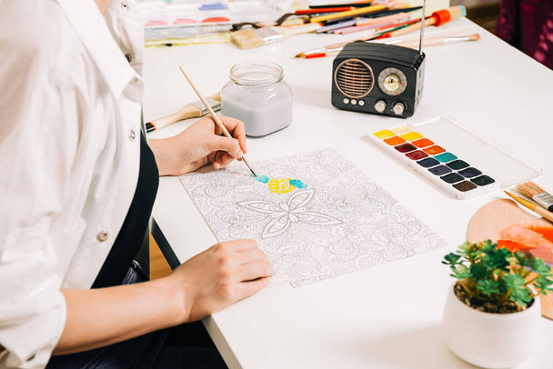Young woman coloring page antistress at table indoors, mental wellbeing and art therapy. Woman paints a sketch, meditative process of coloring pages. - Photo, Image