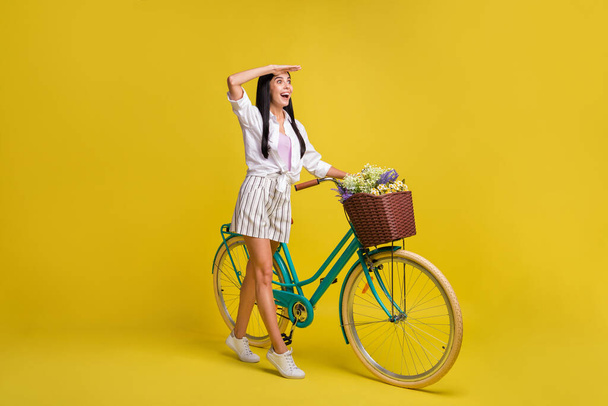 Full size profile side photo of brunette girl look empty space far wear casual outfit hold bike isolated on yellow color background - Foto, Imagen