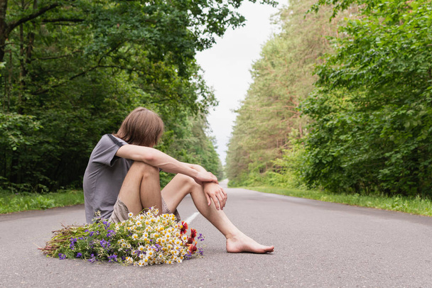 A young guy with flowers is sitting barefoot on the road in a dense forest - Foto, Imagem