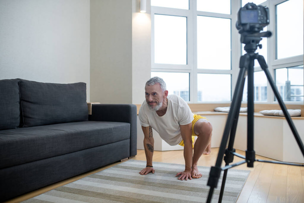 Un hombre maduro en camiseta blanca grabando un tutorial de yoga en línea - Foto, Imagen